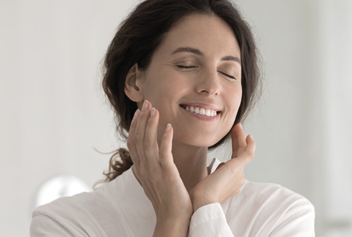 Mujer sonriente con ojos cerrados acariciando su rostro hidratado, reflejando una rutina de cuidado de la piel y bienestar.